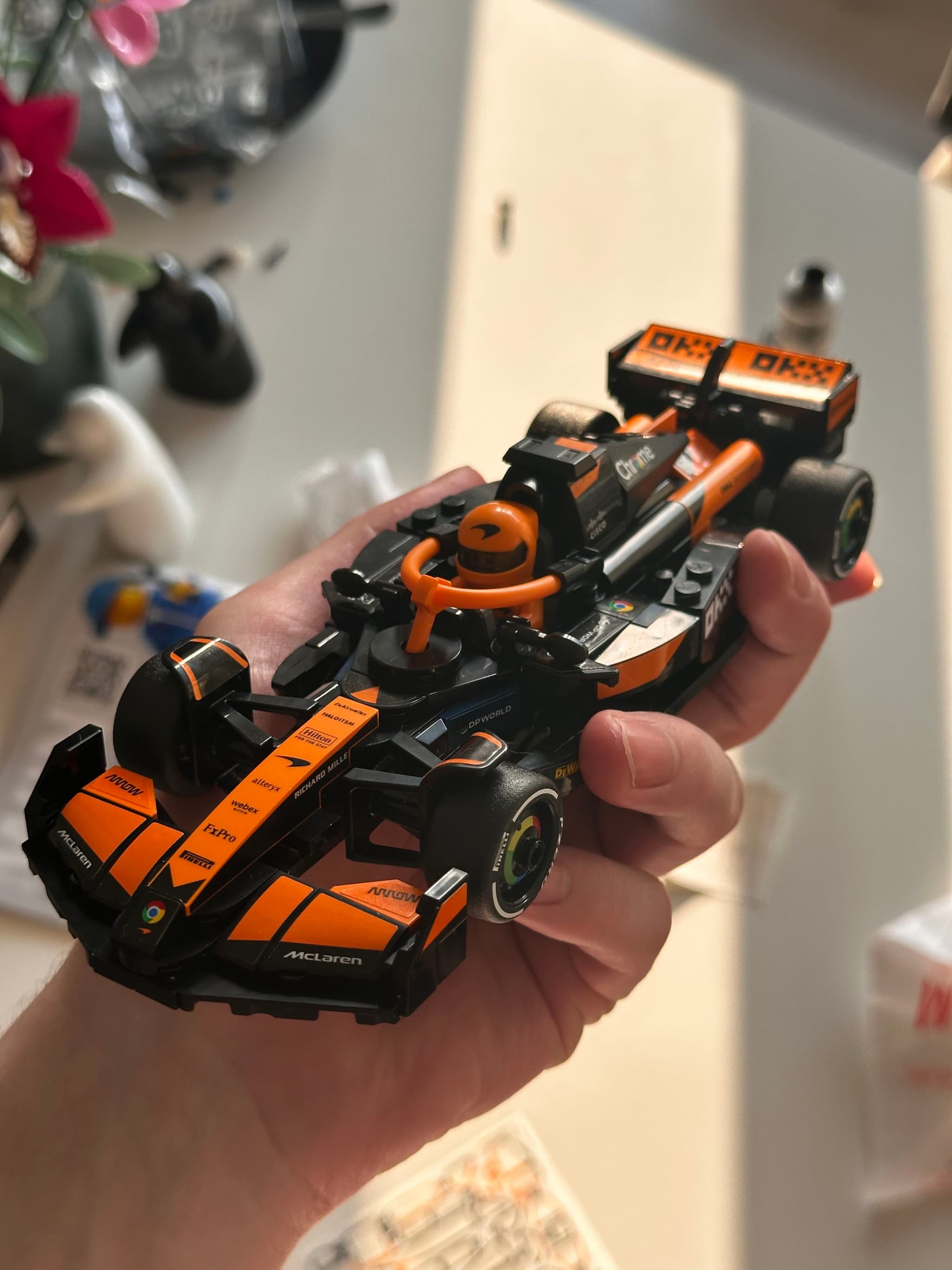 A hand holding a small, detailed model of an orange and black race car, with a blurred background