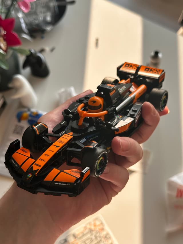 A hand holding a small, detailed model of an orange and black race car, with a blurred background