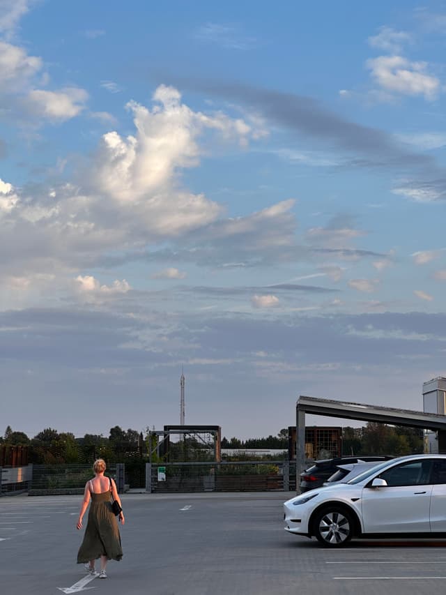 A person walks across a parking lot with a few cars parked, under a partly cloudy sky