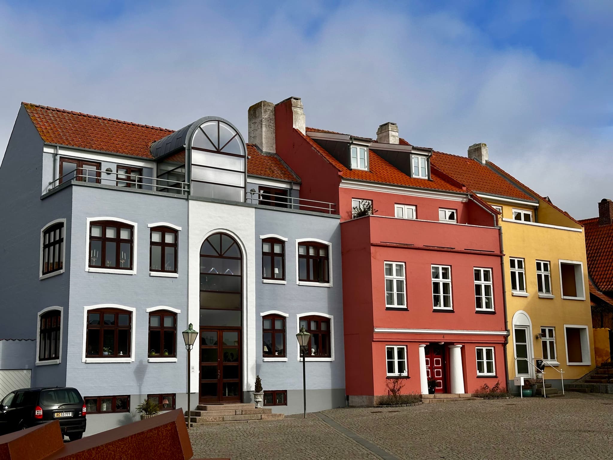 Colorful row of European-style buildings with red, pink, and yellow facades, featuring arched windows and gabled roofs under a partly cloudy sky