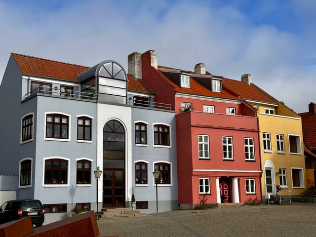 Colorful row of European-style buildings with red, pink, and yellow facades, featuring arched windows and gabled roofs under a partly cloudy sky