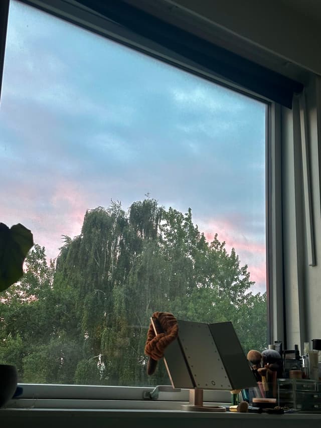 A window view of a tree with a colorful sky in the background, featuring a desk with a tablet and various small items on the windowsill