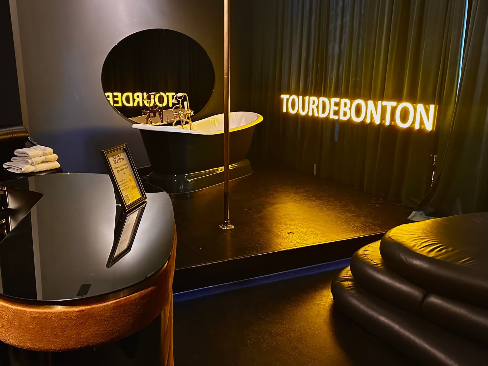 A dimly lit room with a modern, luxurious interior featuring a bathtub, a pole, and neon signs reading TOURDEBONTON A glossy black table and curved seating are also visible