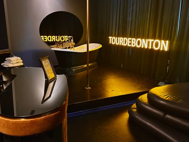 A dimly lit room with a modern, luxurious interior featuring a bathtub, a pole, and neon signs reading TOURDEBONTON A glossy black table and curved seating are also visible
