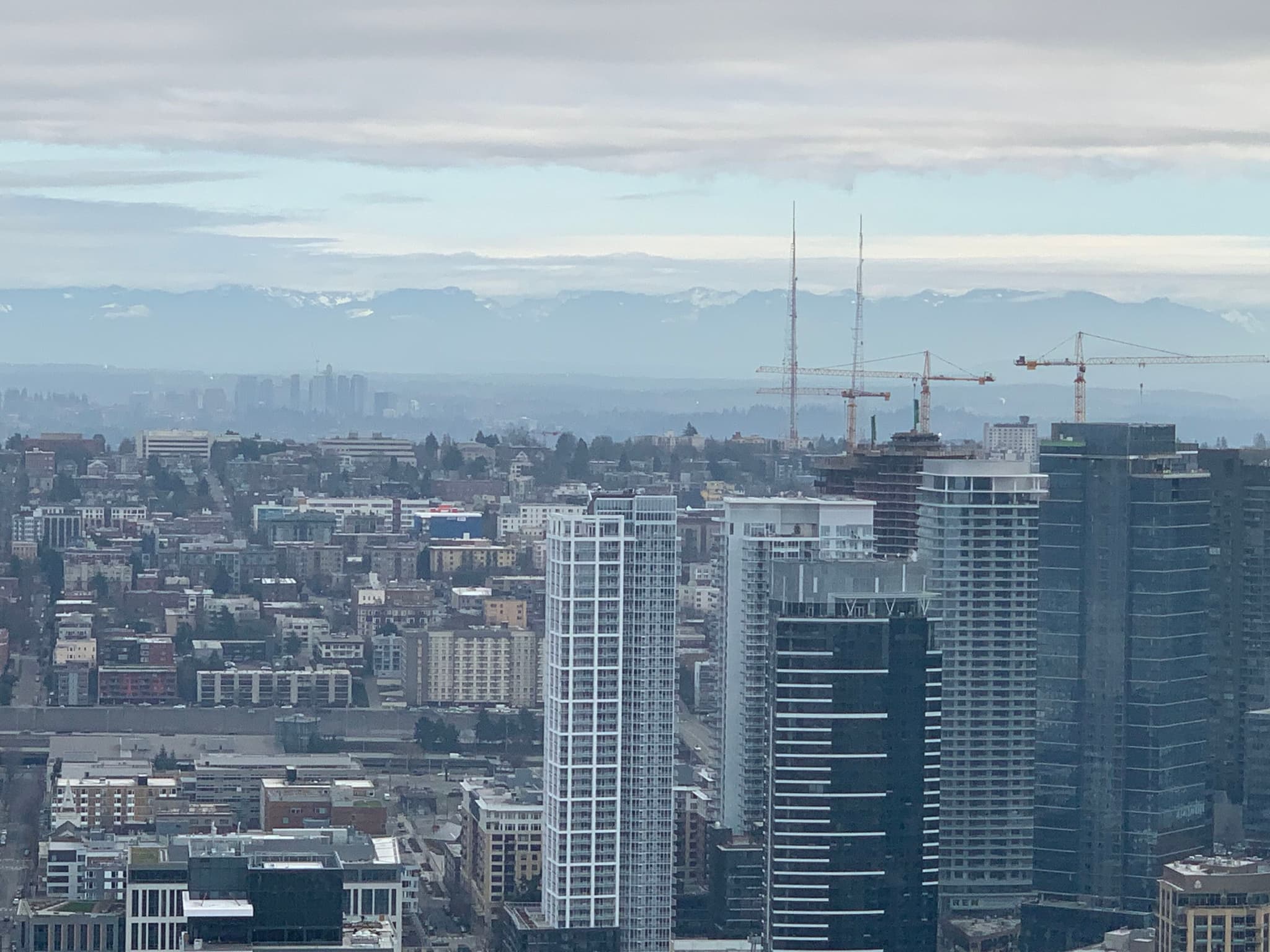 A cityscape featuring tall buildings and cranes under a cloudy sky, with distant mountains visible on the horizon