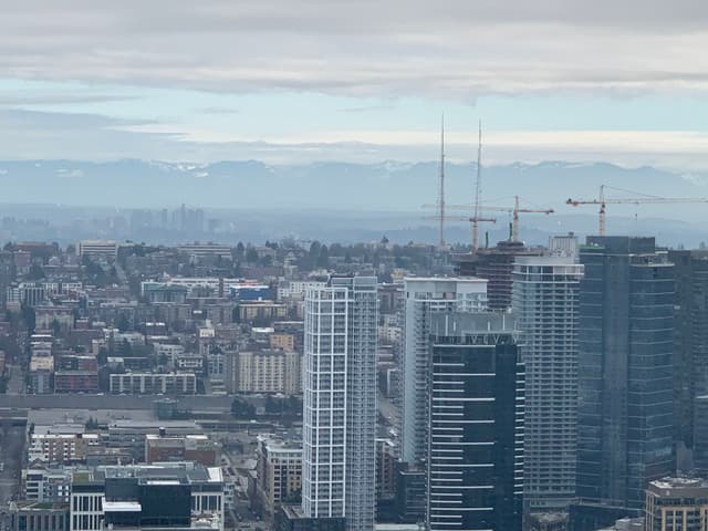 A cityscape featuring tall buildings and cranes under a cloudy sky, with distant mountains visible on the horizon