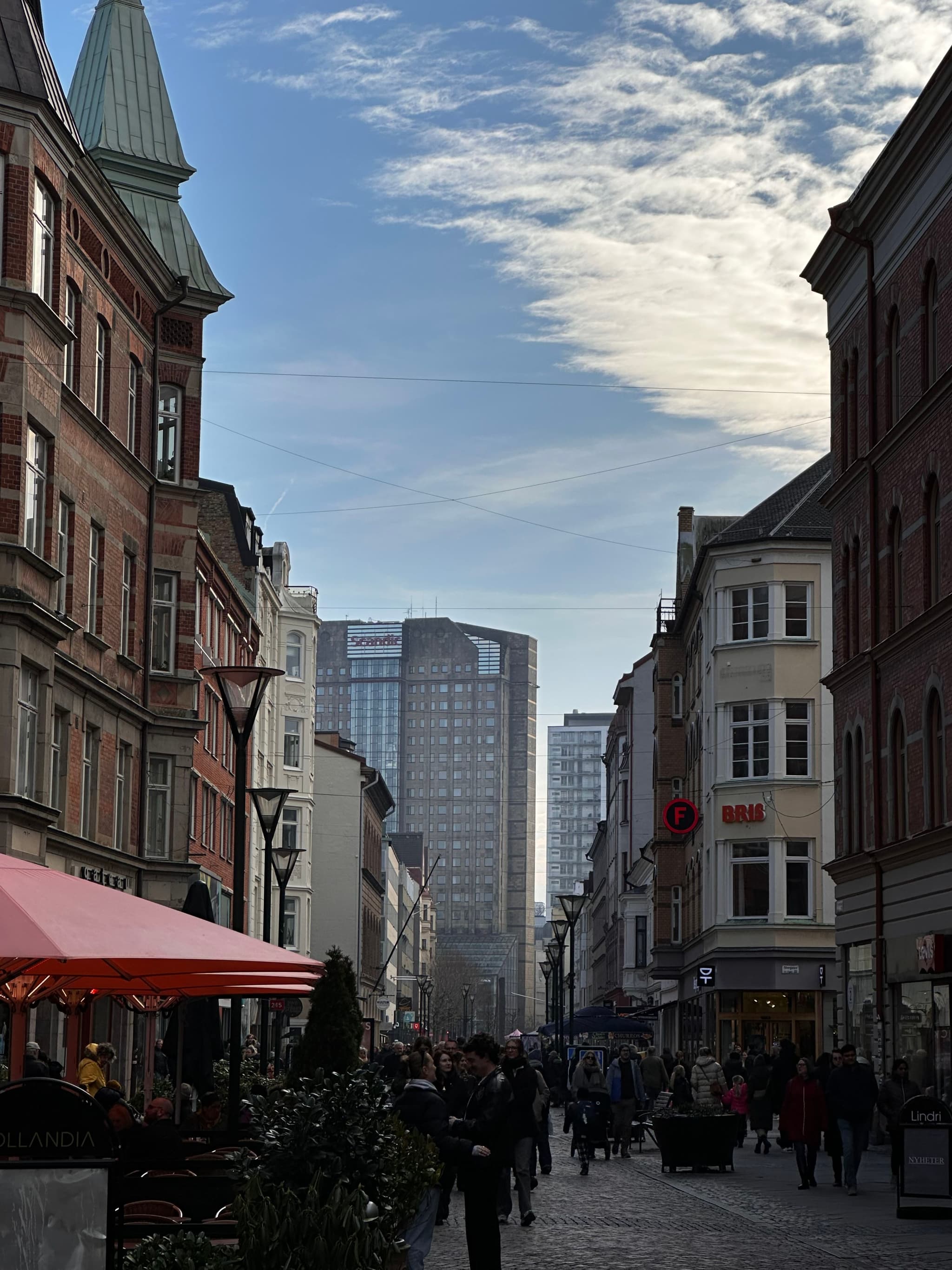 A bustling city street with people walking, flanked by historic buildings on both sides, leading to a modern skyscraper in the background under a partly cloudy sky