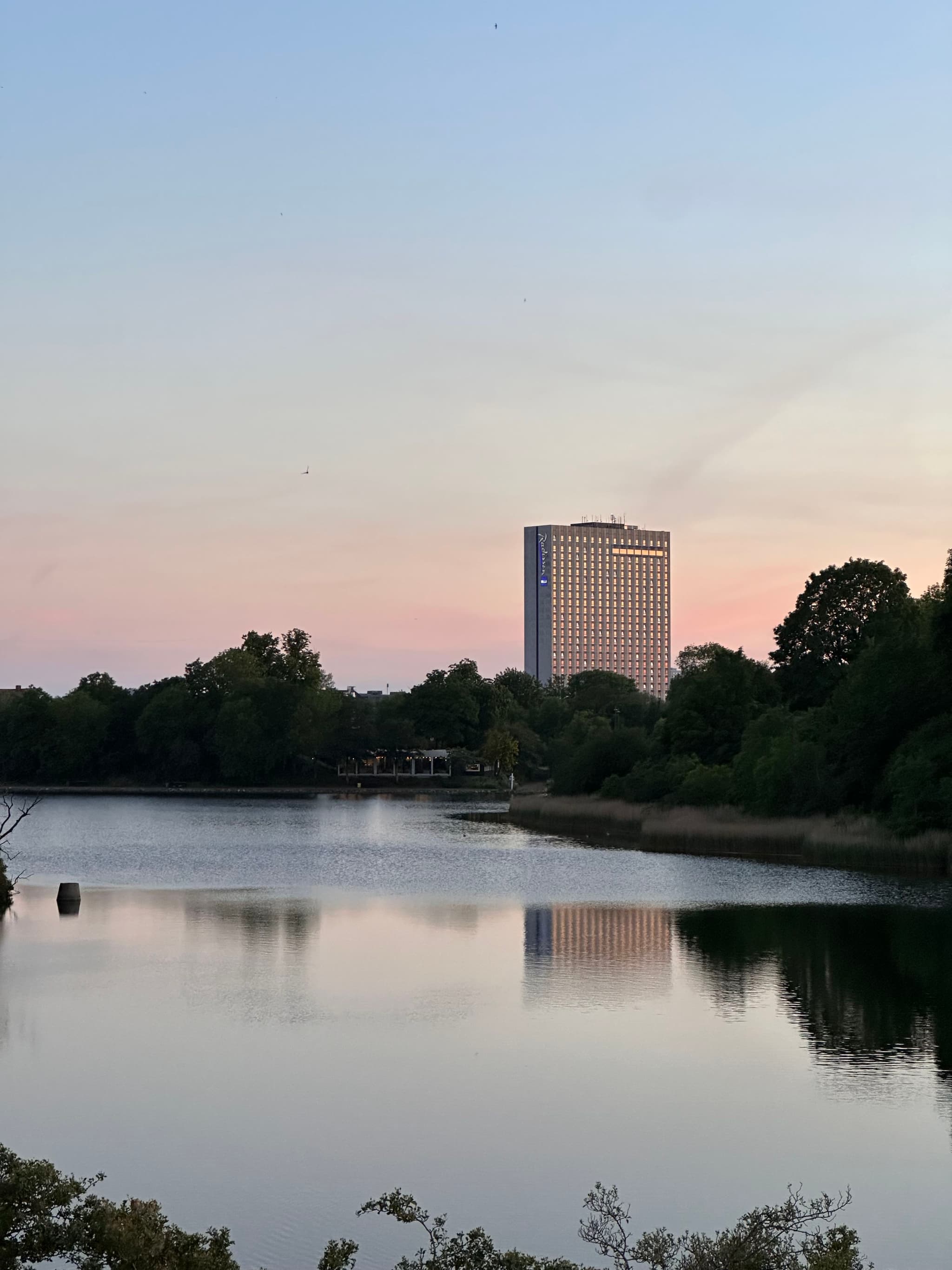 A tranquil lake reflects the surrounding trees and a tall building in the distance, set against a pastel-colored sky at dusk