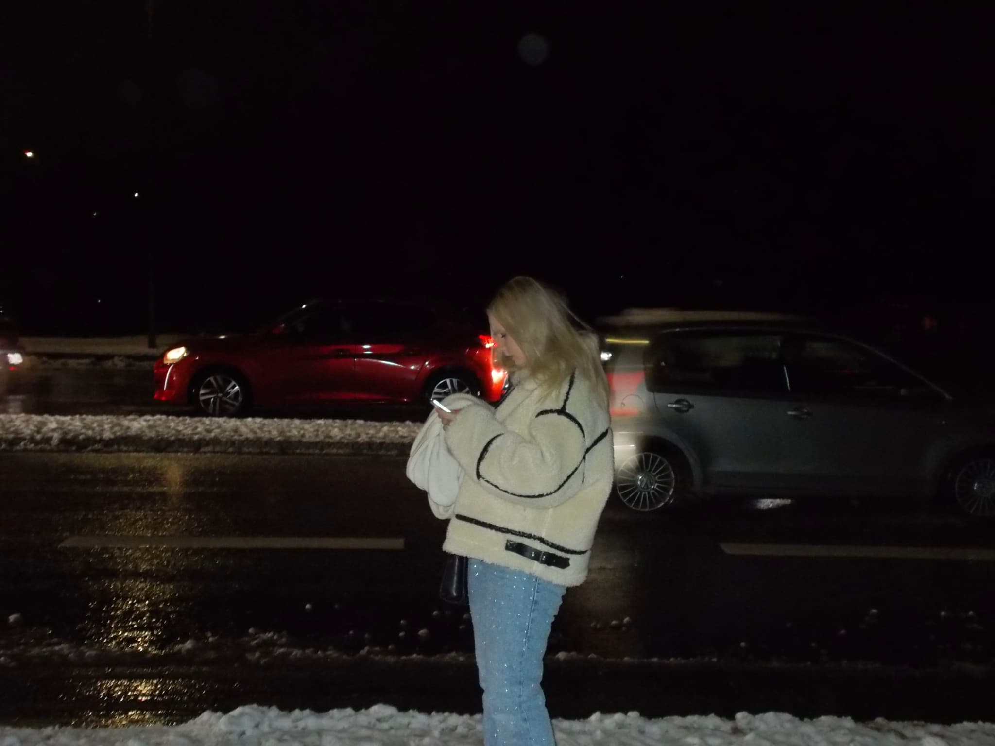 A person in a light-colored jacket stands on a snowy sidewalk at night, looking at a device, with cars passing by on a wet road in the background