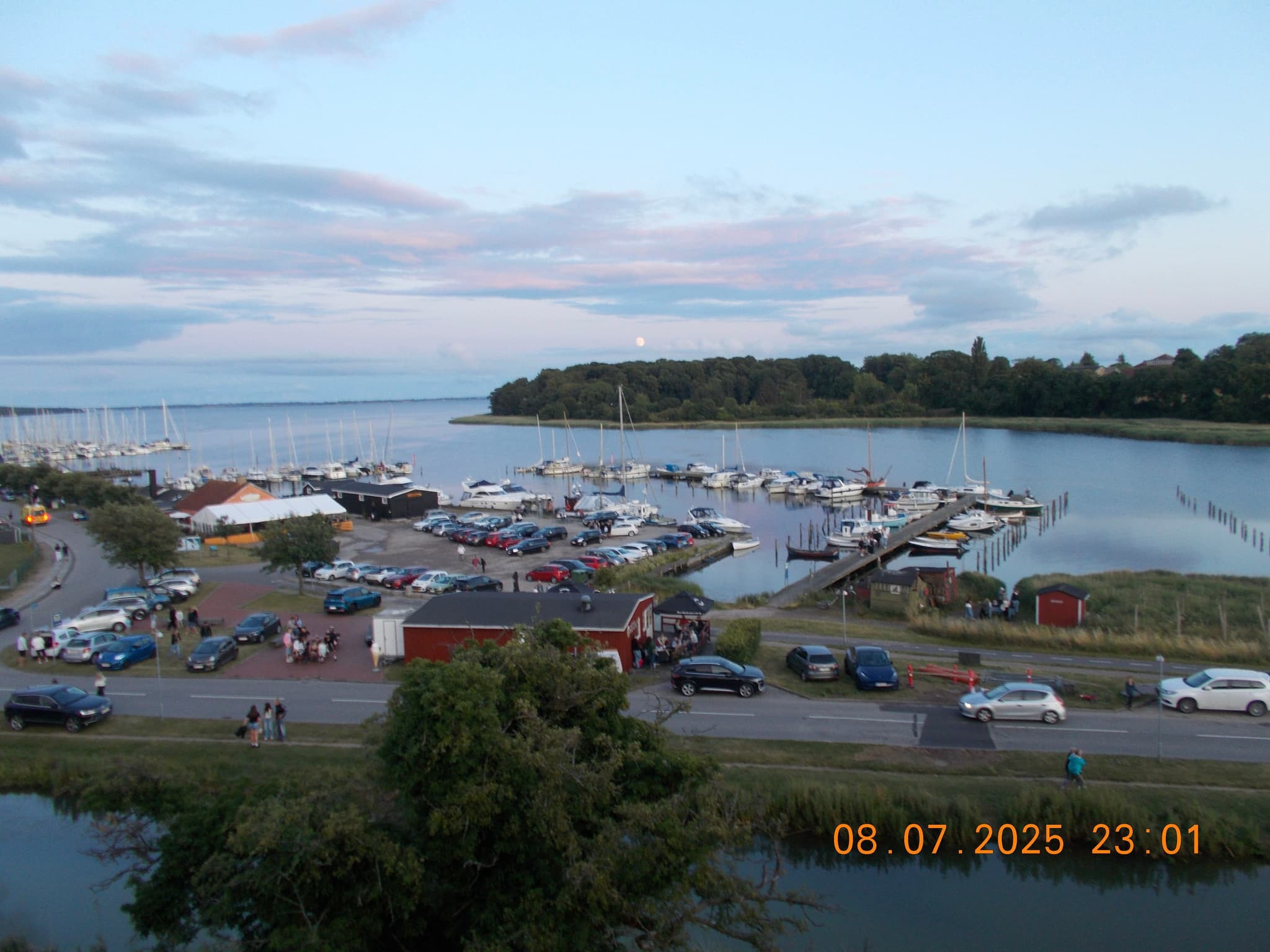 A marina with numerous boats docked, surrounded by parked cars and small buildings, set against a backdrop of calm water and a tree-lined horizon under a partly cloudy sky