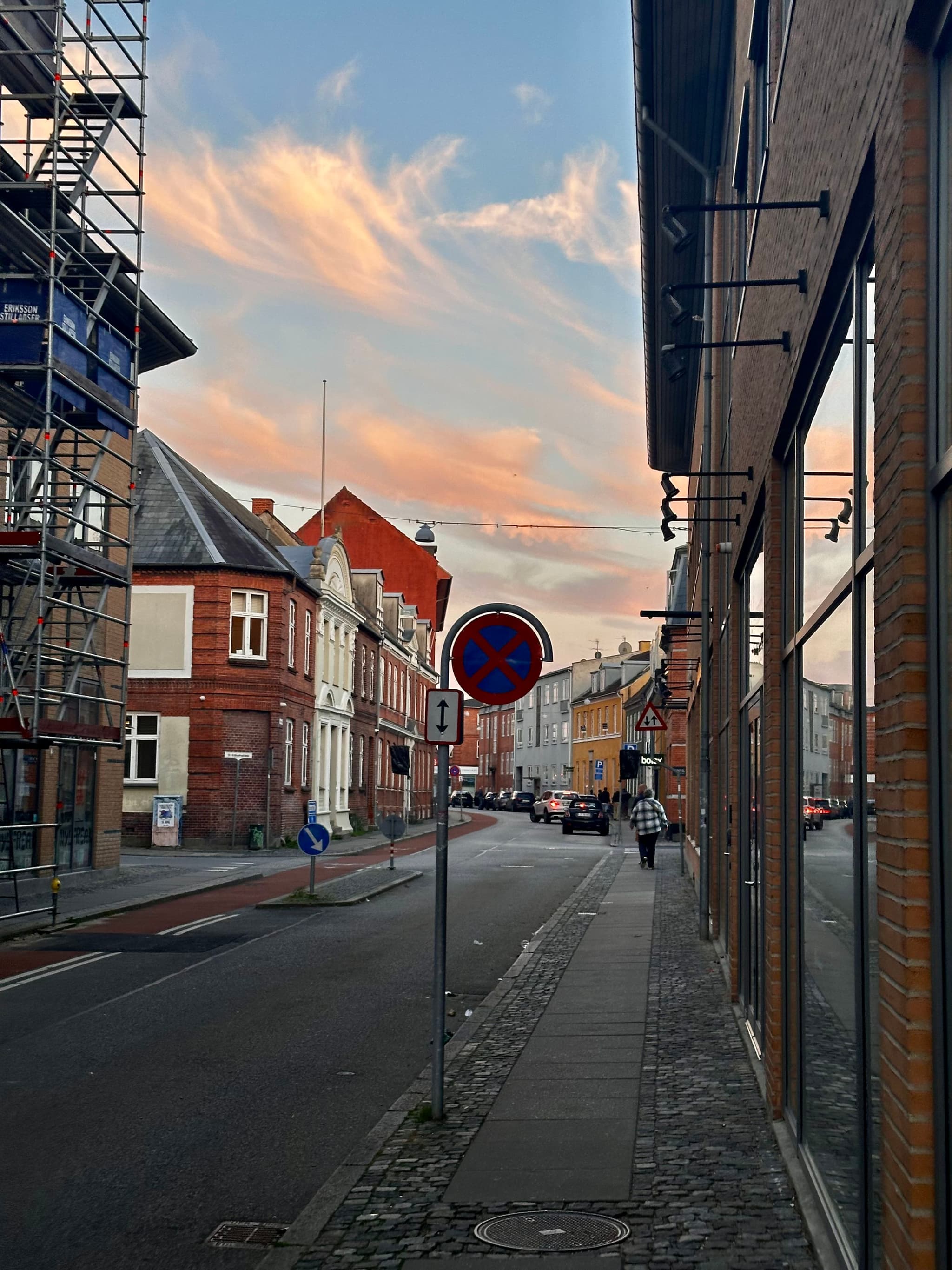 A narrow street lined with brick buildings, featuring a no parking sign, scaffolding on the left, and a few pedestrians. The sky is partly cloudy with a warm sunset glow