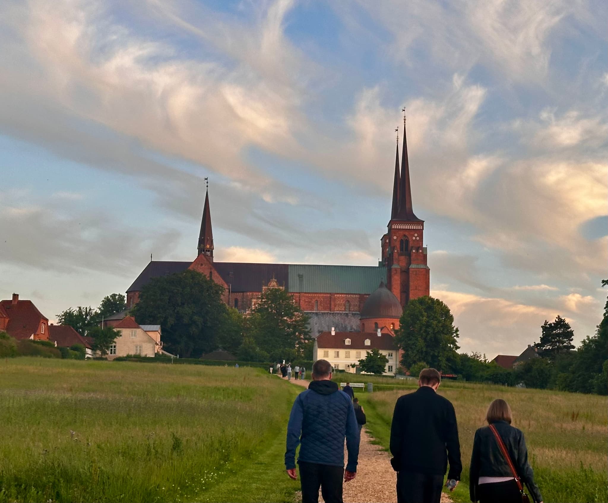 Three people walking on a path towards a large cathedral with two tall spires, surrounded by greenery and under a sky with scattered clouds