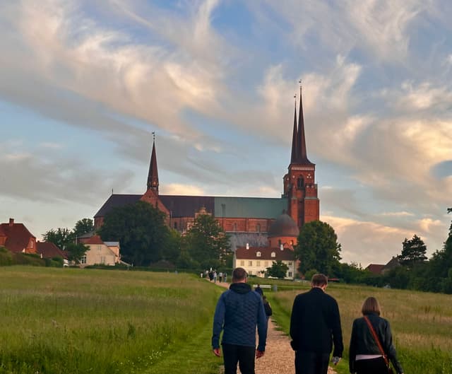 Three people walking on a path towards a large cathedral with two tall spires, surrounded by greenery and under a sky with scattered clouds