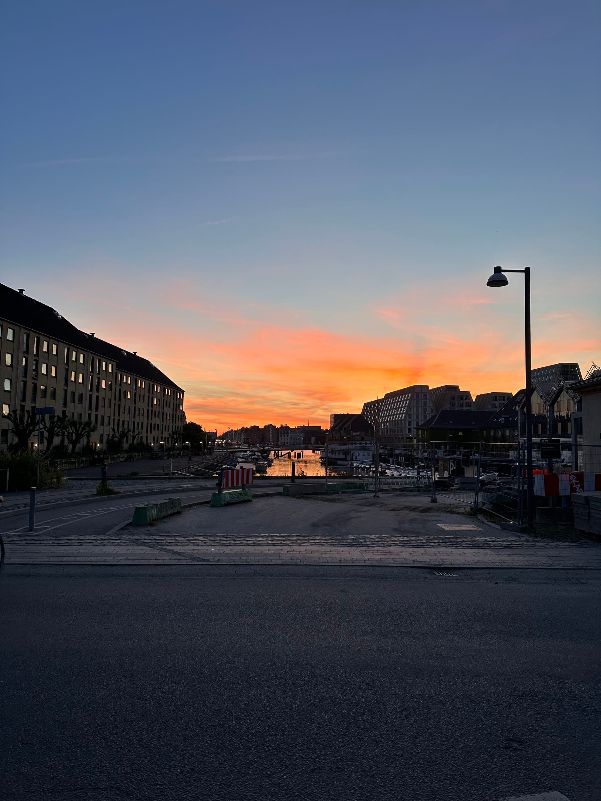 A sunset over a canal flanked by buildings, with a streetlamp silhouetted against the colorful sky