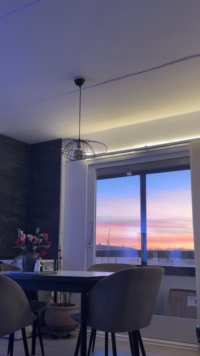 A modern dining area with a table and chairs is illuminated by a ceiling light. A vase of flowers sits on the table, and a large window reveals a colorful sunset outside
