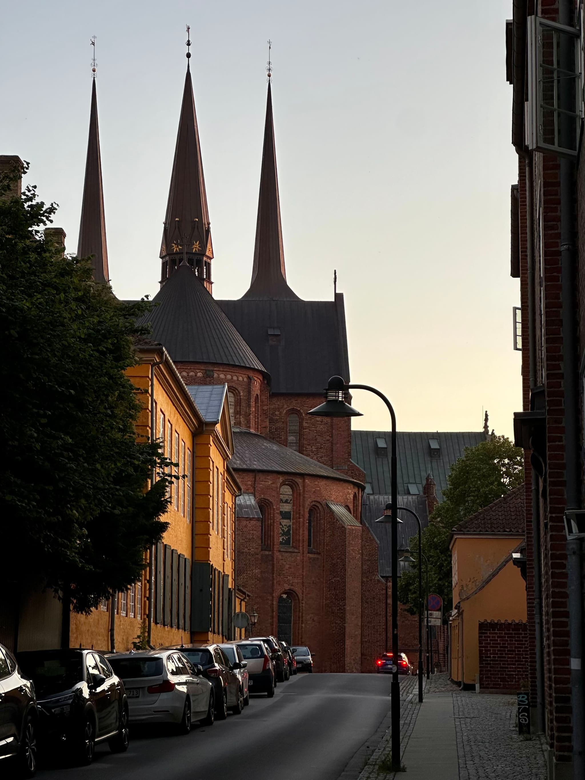 A narrow street lined with parked cars and buildings leads to a church with tall spires in the background, under a clear sky