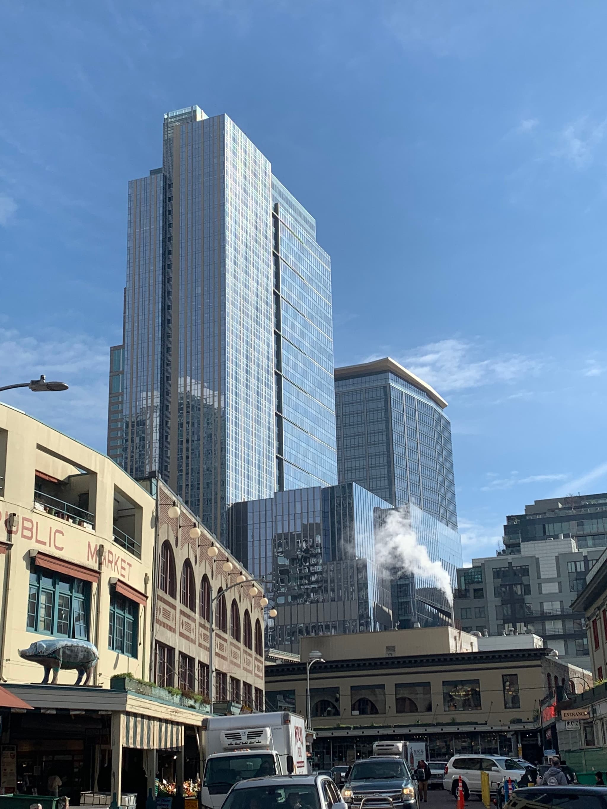A cityscape featuring tall modern skyscrapers with reflective glass facades, surrounded by older, low-rise buildings. The sky is clear and blue, and there is light traffic on the street below