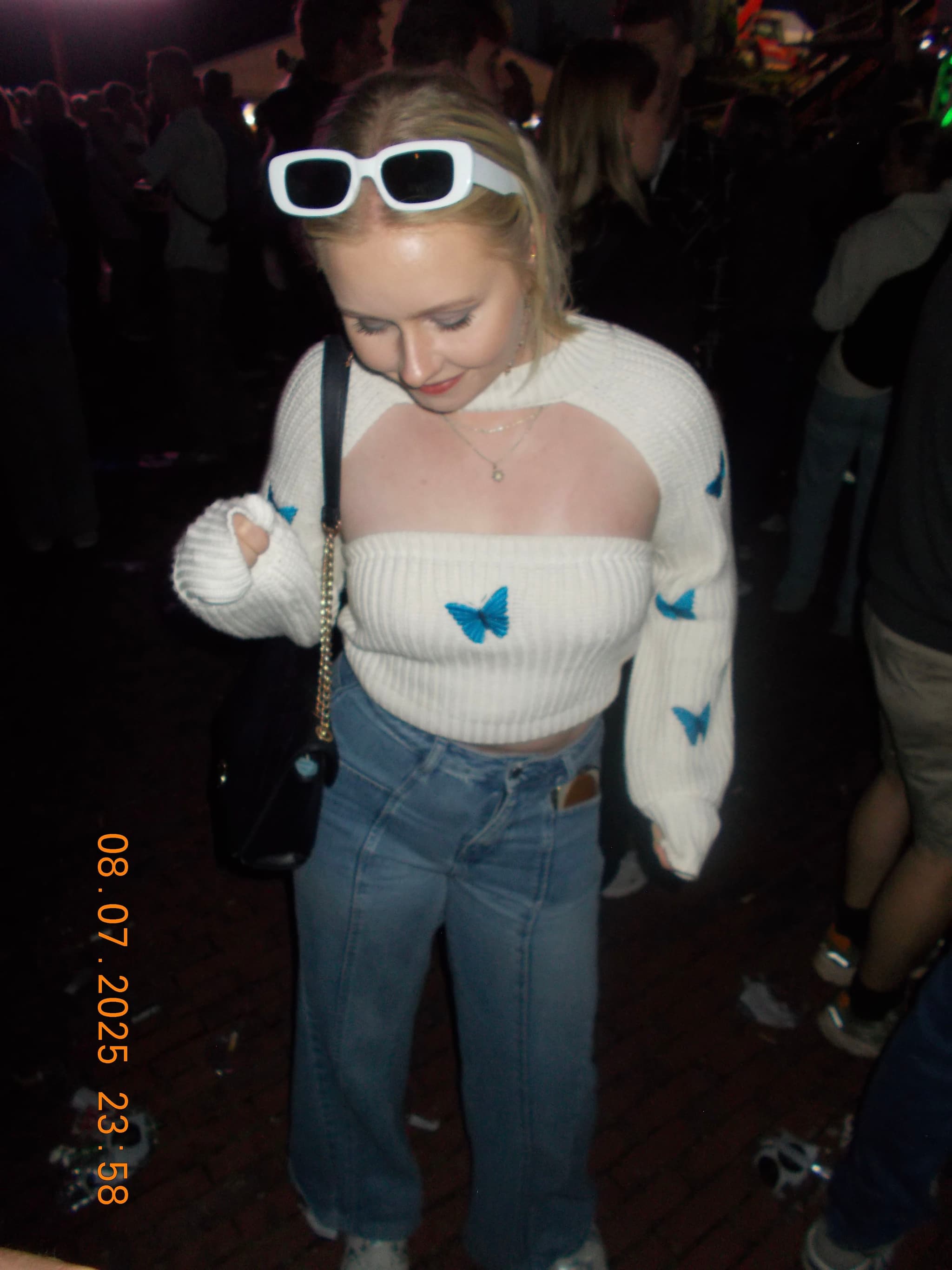 A person wearing a white sweater with blue butterfly designs, blue jeans, and white sunglasses on their head, standing in a dimly lit, crowded area