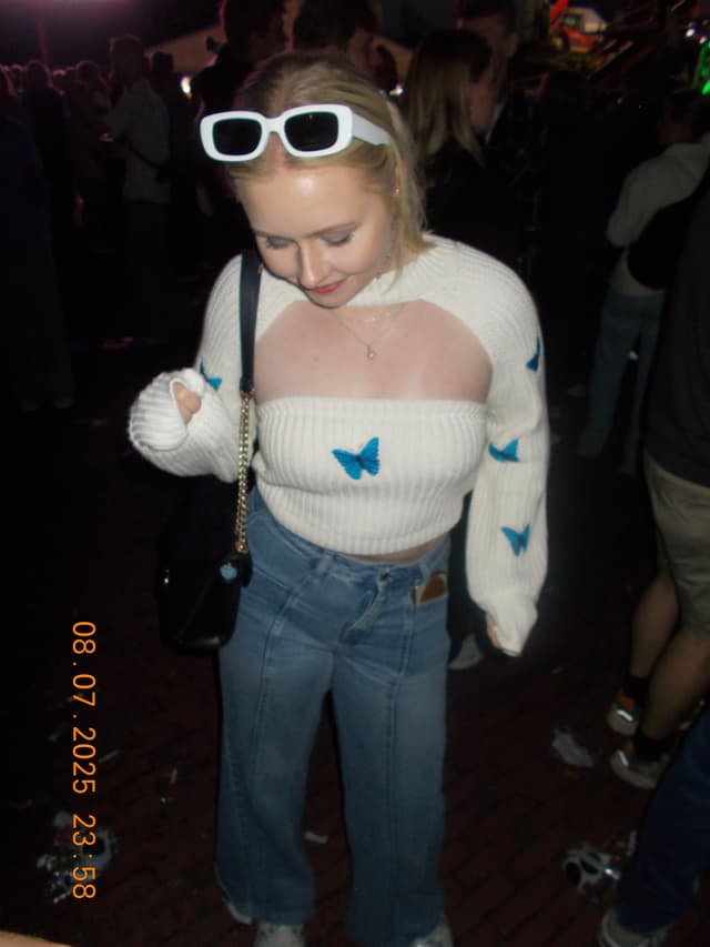 A person wearing a white sweater with blue butterfly designs, blue jeans, and white sunglasses on their head, standing in a dimly lit, crowded area