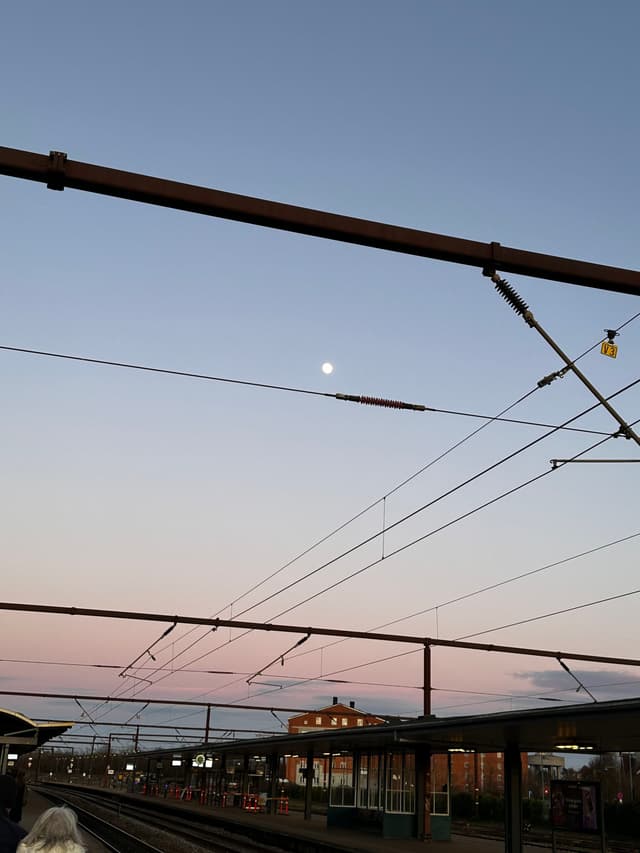 A railway station at dusk with overhead power lines, a visible moon in the sky, and a building in the background